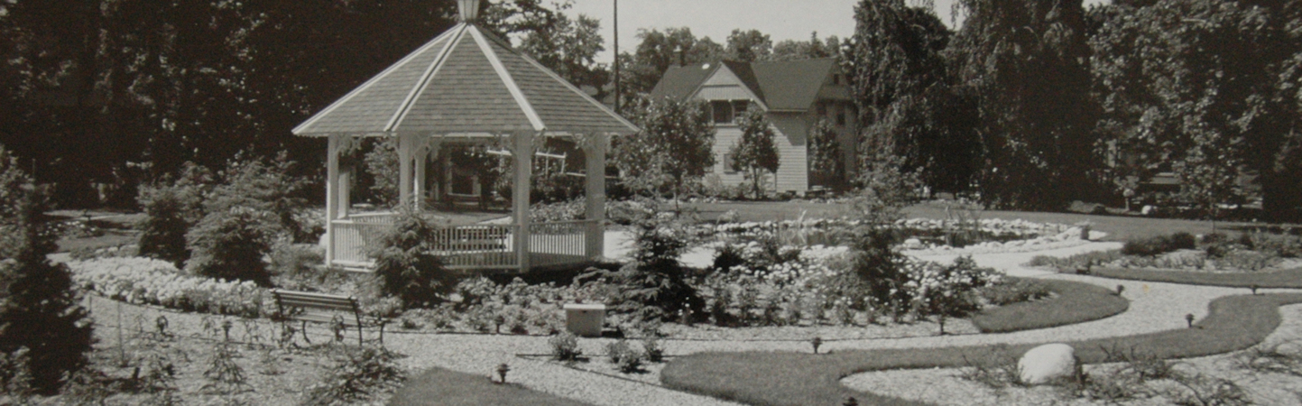 Black and white photo of wooden gazebo in manicured garden with bed of annuals and crushed stone pathways.