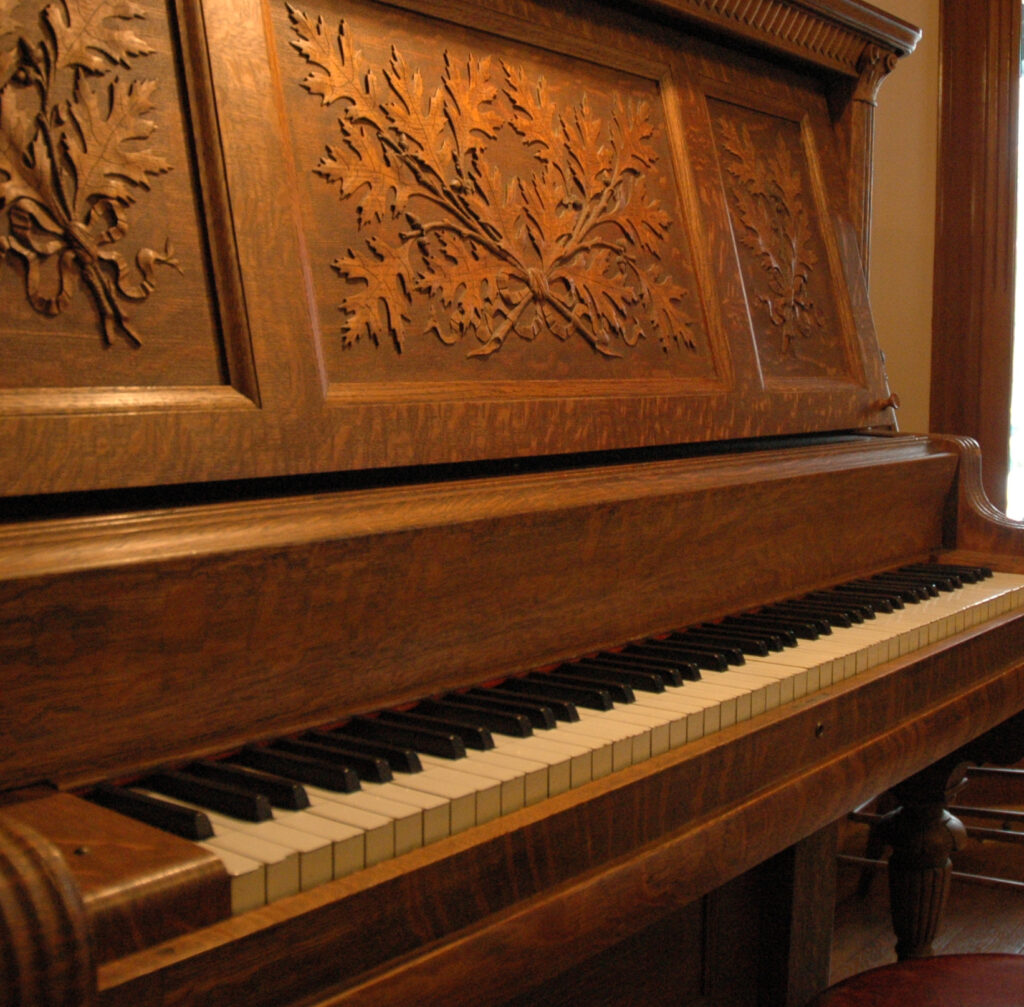 Angled view of keys of a 1908 Upright Piano with ornate wooden carving in a parlor with a window.